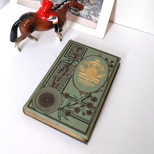 May include: A vintage equestrian figurine of a rider in a red coat on a brown horse. Beside it is a green book with gold lettering that reads "Pilgrim's Progress". A vintage illustration with the word "BEST" is in the background.