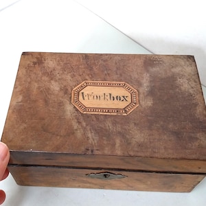 Peut inclure: Une boîte rectangulaire en bois avec une finition brun foncé et une texture patinée. Le couvercle présente une incrustation octogonale centrale avec le mot Workbox imprimé en noir. Une petite plaque de serrure en métal est centrée sur la face avant.