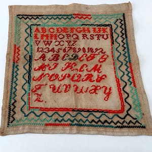 以下が含まれることがあります： 赤い糸で刺繍された文字と数字が特徴のヴィンテージサンプラー。サンプラーはベージュの背景に赤、緑、黒の装飾的なボーダーがあります。テキストにはアルファベットと数字が含まれています。