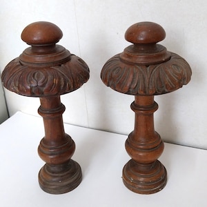 May include: Two antique wooden candle holders. Each holder has a round base, a turned stem, and a carved, flared top with a small, rounded finial. The wood has a warm, brown tone, and the pieces appear to be vintage.