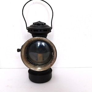 May include: Antique black metal lantern with a clear glass lens and a silver-colored rim. The lantern has a handle and a round base. The design is reminiscent of vintage railway or carriage lamps.