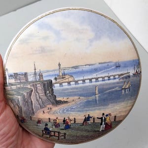 May include: A circular antique plate with a coastal scene, including a pier, lighthouse, and figures in period dress. The plate has a gold rim and a detailed, colourful illustration of a seaside town.