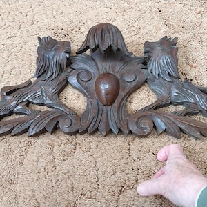 May include: A dark brown, carved wooden architectural element. It features two stylized lion figures either side of a central floral design. The piece has intricate leaf and scroll details, and is likely a decorative accent.
