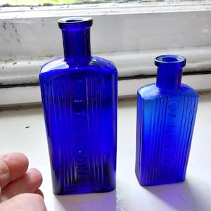 May include: Two antique cobalt blue glass bottles. The bottles have a rectangular shape with vertical ridges and a small neck. One bottle is taller than the other. The bottles are sitting on a white surface.