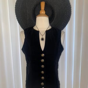 May include: A black velvet vest featuring a deep V-neckline and six ornate metallic buttons down the front. The vest is displayed on a mannequin wearing a wide-brimmed black straw hat and a silver-toned pendant necklace with a dark oval stone.