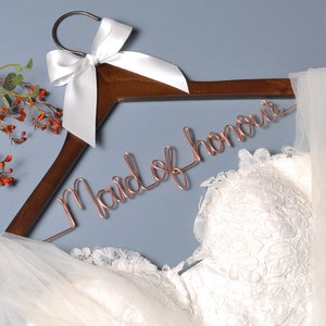 K&ouml;nnte beinhalten: Ein Holz Kleiderb&uuml;gel mit einer wei&szlig;en Satin Schleife und einer ros&eacute;goldenen Drahtinschrift mit der Aufschrift "Maid of Honor". Ein wei&szlig;er T&uuml;ll Schleier und ein wei&szlig;es Spitzen Brautkleid sind &uuml;ber den Kleiderb&uuml;gel gelegt.