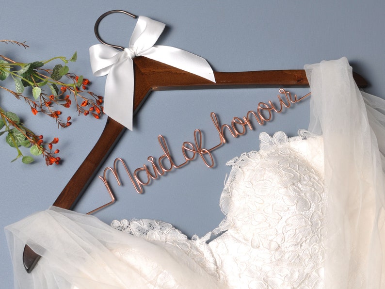 K&ouml;nnte beinhalten: Ein Holz Kleiderb&uuml;gel mit einer wei&szlig;en Satin Schleife und einer ros&eacute;goldenen Drahtinschrift mit der Aufschrift "Maid of Honor". Ein wei&szlig;er T&uuml;ll Schleier und ein wei&szlig;es Spitzen Brautkleid sind &uuml;ber den Kleiderb&uuml;gel gelegt.