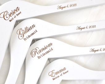 Könnte beinhalten: Vier weiße Holzbügel mit goldener Beschriftung. Die Bügel sind mit den Namen "Cesca", "Gillian", "Ransom" und "Emma" sowie ihren entsprechenden Titeln "Maid of Honor", "Bridesmaid", "Bridesmaid" und "Matron of Honor" personalisiert. Jeder Bügel enthält auch das Datum "August 4, 2023".