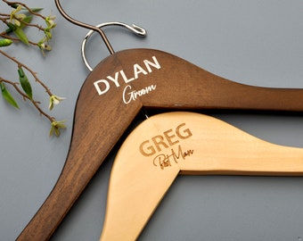 Könnte beinhalten: Zwei Holzbügel mit graviertem Text. Der obere Bügel ist dunkelbraun mit dem Text "DYLAN Groom". Der untere Bügel ist hellbraun mit dem Text "GREG Best Man".