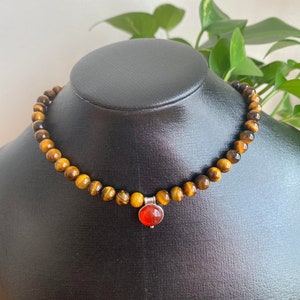 May include: A beaded necklace with tiger's eye stones and a pendant with a round, orange gemstone. The necklace is displayed on a black mannequin. The tiger's eye beads are approximately 0.8 cm in diameter.