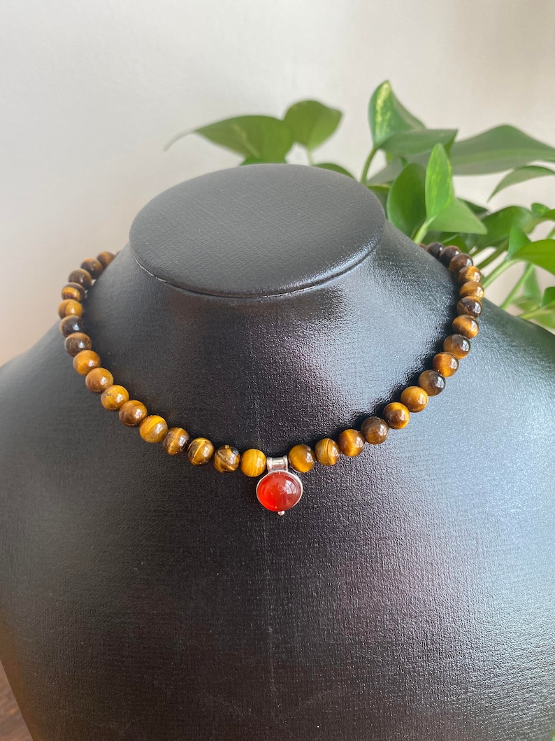 May include: A beaded necklace with tiger's eye stones and a pendant with a round, orange gemstone. The necklace is displayed on a black mannequin. The tiger's eye beads are approximately 0.8 cm in diameter.