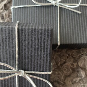 May include: Two black gift boxes with vertical striped patterns, tied with silver string bows. The boxes are on a soft, textured surface. These boxes are likely for presents or small items.