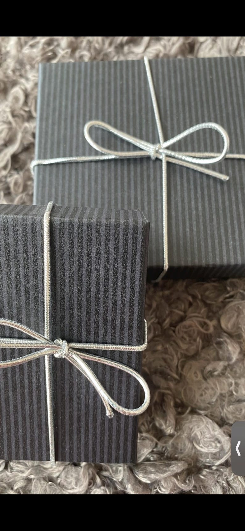 May include: Two black gift boxes with vertical striped patterns, tied with silver string bows. The boxes are on a soft, textured surface. These boxes are likely for presents or small items.