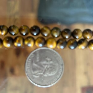 May include: Two strands of polished tiger eye gemstone beads, each bead approximately 0.8 cm in diameter, are displayed next to a US quarter coin. The beads have a warm, golden-brown colour with dark brown streaks.