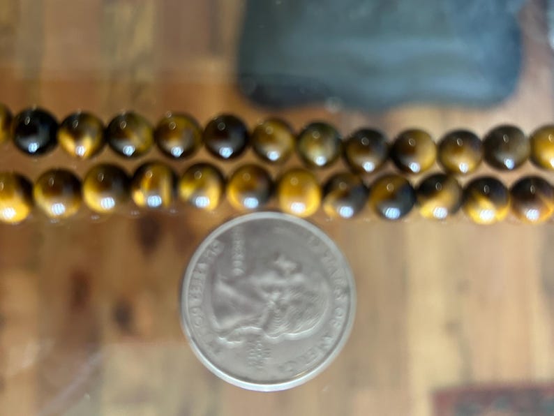 May include: Two strands of polished tiger eye gemstone beads, each bead approximately 0.8 cm in diameter, are displayed next to a US quarter coin. The beads have a warm, golden-brown colour with dark brown streaks.