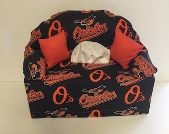 May include: A black and orange tissue box cover shaped like a couch with the Baltimore Orioles logo and the words "O's Orioles" repeated on the fabric.