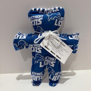 May include: A handmade doll in the shape of a person, made from blue fabric printed with the Detroit Lions logo. The doll has a white tag that reads "Damn It Doll" with instructions. The doll is tied with white string.