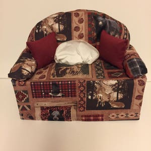 May include: A tissue box cover shaped like a couch with a brown, red, and green plaid pattern. The couch has two red pillows and a white tissue box inside.