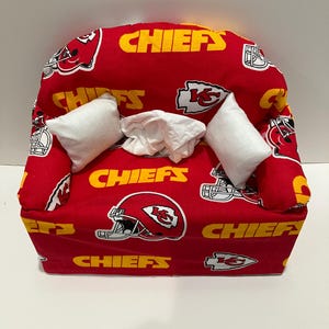 May include: Red and yellow tissue box cover shaped like a couch with two white pillows. The cover has the Kansas City Chiefs logo and the word "Chiefs" repeated on it.