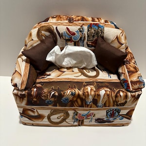 May include: A fabric tissue box cover with a Western theme. The cover features a repeating pattern of horses, saddles, cowboy boots, and ropes in shades of brown, tan, and blue. Two small brown pillows flank the tissue opening.
