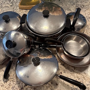 May include: A collection of stainless steel cookware, including pots, pans, and lids. The pots and pans have black handles and knobs. The lids are domed and reflect the surrounding environment.