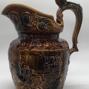 May include: A brown ceramic pitcher with a detailed design of a horse and rider, flowers, and a building. The pitcher has a handle shaped like a horse's head.
