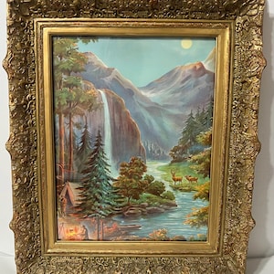 May include: A framed vintage print of a scenic landscape with a waterfall, mountains, trees, a river, and deer. The print is in a gold ornate frame.