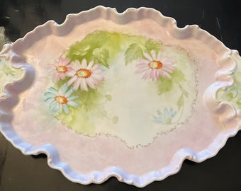 May include: A vintage porcelain serving tray with a pink background and a floral design featuring pink and blue daisies. The tray has a scalloped edge and two handles.