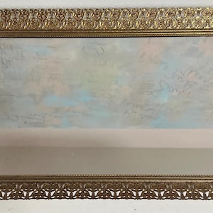 May include: A gold-colored ornate picture frame with a decorative filigree design. The frame is empty and has a light blue and pink background.