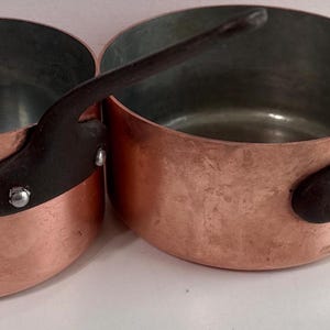 May include: Two copper saucepans with dark brown handles. The pans have a rounded shape with a slightly flared rim. The interior of the pans is a darker metal, and the exterior is a shiny copper color. The handles are attached with rivets.
