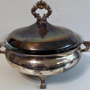 May include: A silver-plated serving dish with a glass insert and lid. The dish has ornate handles and a decorative handle on the lid. The dish sits on three decorative feet. The lid has a reflective surface.