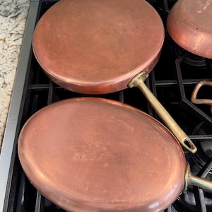 May include: Two round copper pans with smooth, metallic surfaces and brass handles. The pans are positioned with their flat bases facing upward, showing a warm, reddish-brown tone and a polished finish. Each pan features a sturdy, riveted brass handle.