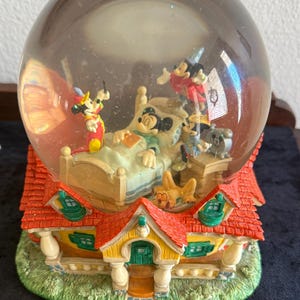 Könnte beinhalten: Eine Disney-Schneekugel mit Mickey Mouse, die mit anderen Figuren in einem Bett schläft, auf einem Haus-Sockel. Ein Holzlineal zeigt, dass die Basis etwa 17,8 cm breit ist. Die Kugel enthält Flüssigkeit und Glitzerpartikel.