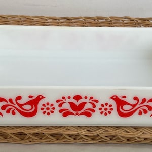 May include: White rectangular baking dish with red floral and bird design. The dish is sitting on a woven wicker placemat. The dish has a rectangular shape with slightly flared sides and handles.
