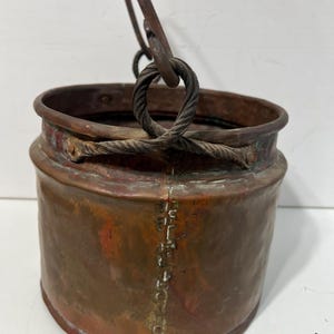 May include: A copper bucket with a twisted wire handle. The bucket has a worn, aged patina and is made of hammered copper.