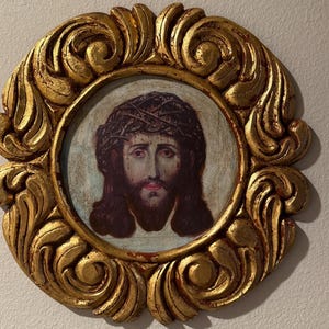 May include: A framed religious icon featuring a portrait of Jesus Christ wearing a crown of thorns. The portrait is set within a gold-colored, ornate, oval frame with a detailed, swirling design. The artwork has a vintage aesthetic.