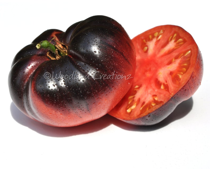 May include: A close-up of a dark purple and red heirloom tomato, with water droplets on the surface. One tomato is whole, and the other is cut in half, revealing the red interior. The image includes the text "Woodland Creationsz".