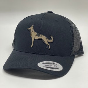 May include: Black trucker hat with a black and tan dog silhouette on the front. The hat has a black mesh back and a black brim. The hat has a label on the back that says "The Authentic Yupoong Snapback Yupoong Since 1974".