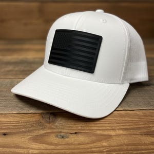 May include: White trucker hat with a black American flag patch. The hat features a curved brim and a mesh back for breathability. The flag patch is rectangular and prominently displayed on the front.