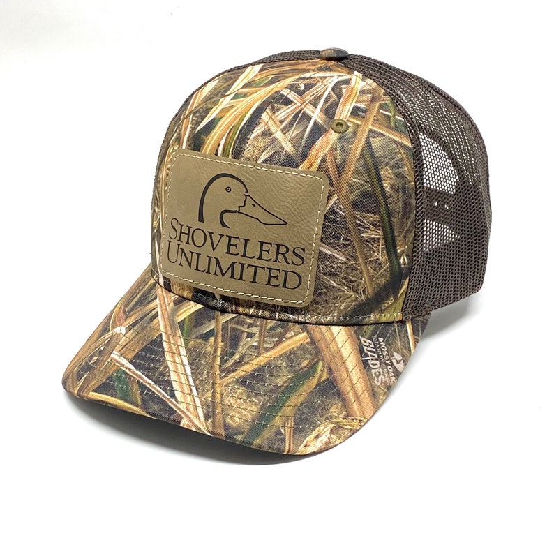 Puede incluir: Un gorro de camionero de camuflaje marr&oacute;n y verde con un parche de cuero marr&oacute;n que dice "Shovelers Unlimited" y una silueta de pato marr&oacute;n. El gorro tiene una parte trasera de malla marr&oacute;n.