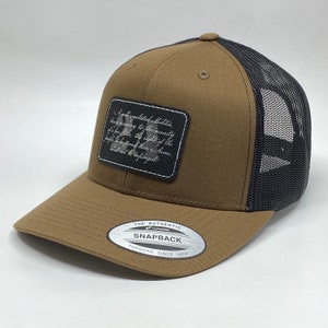 Op de afbeelding: Een bruine en zwarte baseballpet met een mesh achterkant. De pet heeft een patch aan de voorkant met de tekst "A well regulated Militia, being necessary to the security of a free State, the right of the people to keep and bear Arms, shall not be infringed." De pet heeft ook een zilveren sticker aan de zijkant met de tekst "The Authentic Yupoong Snapback Since 1974."