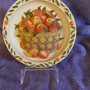 Puede incluir: Un plato de cerámica decorativo con un diseño en relieve de una cesta llena de melocotones. El plato tiene un fondo crema con hojas verdes y una cesta de mimbre marrón.