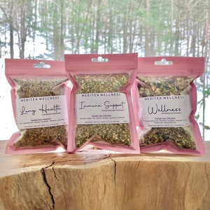 May include: Three pink pouches of herbal tea infusions, labeled "Lung Health," "Immune Support," and "Wellness." Each pouch contains dried herbs and is labeled "Meditea Wellness." The background features a blurred view of a snowy forest.