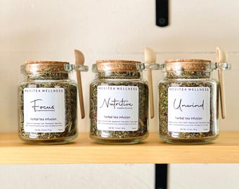 May include: Three glass jars of herbal tea infusions labeled "Focus", "Nutritive", and "Unwind". Each jar has a cork lid and a small wooden spoon attached. The jars are filled with dried herbs and sit on a wooden shelf.