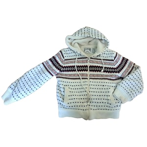 May include: A cream-colored zip-up hooded sweater with a patterned design. The sweater features a white interior lining in the hood and a repeating pattern of small black hearts and burgundy geometric shapes. The sweater has two front pockets.