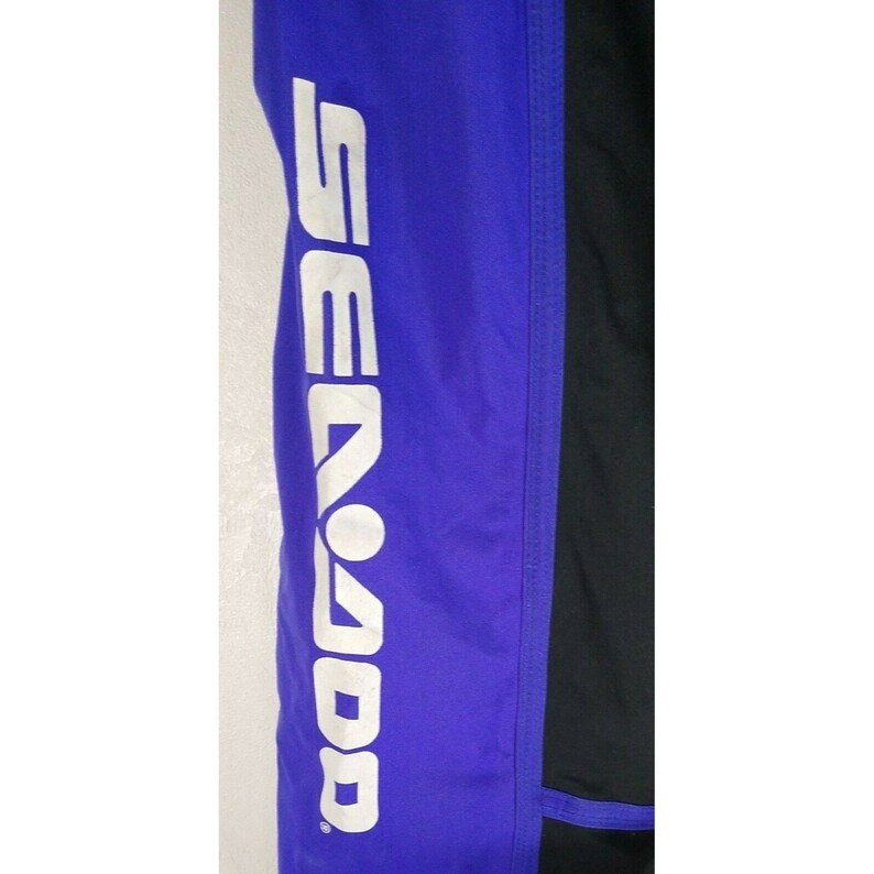 May include: Close-up of a section of a garment with a blue panel and a black panel. The word "SEADOO" is printed vertically in white on the blue panel. The garment appears to be made of a synthetic material.