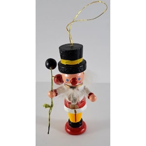 May include: A wooden Christmas ornament depicting a man wearing a black top hat with a yellow band, a white beard, and a red and white jacket. He is holding a stick with a black ball on top.