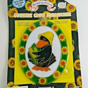 Op de afbeelding: Een gebrandschilderd zonnenvanger met een zwarte raaf die een gele hoed draagt en een kolf maïs vasthoudt. De raaf wordt omringd door een rand van gele zonnebloemen met groene bladeren. De zonnenvanger is ovaalvormig en heeft een zuignap om op te hangen. De verpakking bevat de tekst "Country Crow Suncatcher" en "Hermitage Pottery".