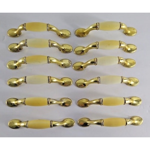 Könnte beinhalten: Zehn goldene Schrankknöpfe mit einem cremefarbenen Keramikeinsatz. Die Knöpfe haben ein schlichtes, elegantes Design.