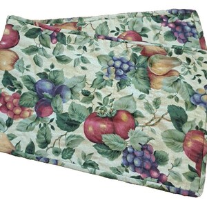 May include: Two quilted placemats with a fruit and foliage pattern. The design features red apples, purple grapes, and green leaves on a cream-colored background. The placemats are rectangular and appear to be made of fabric.
